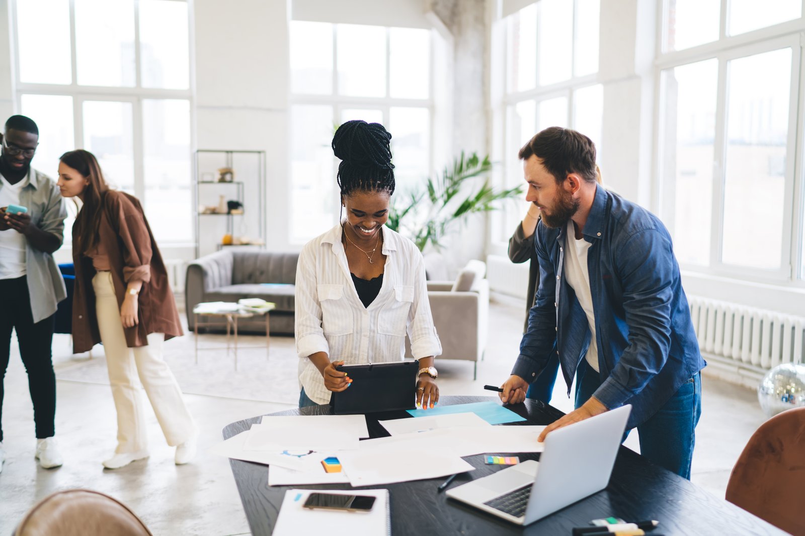 Group of multiracial businesspeople standing at wooden table with papers laptop and smartphone while discussing business startup in modern workshop together