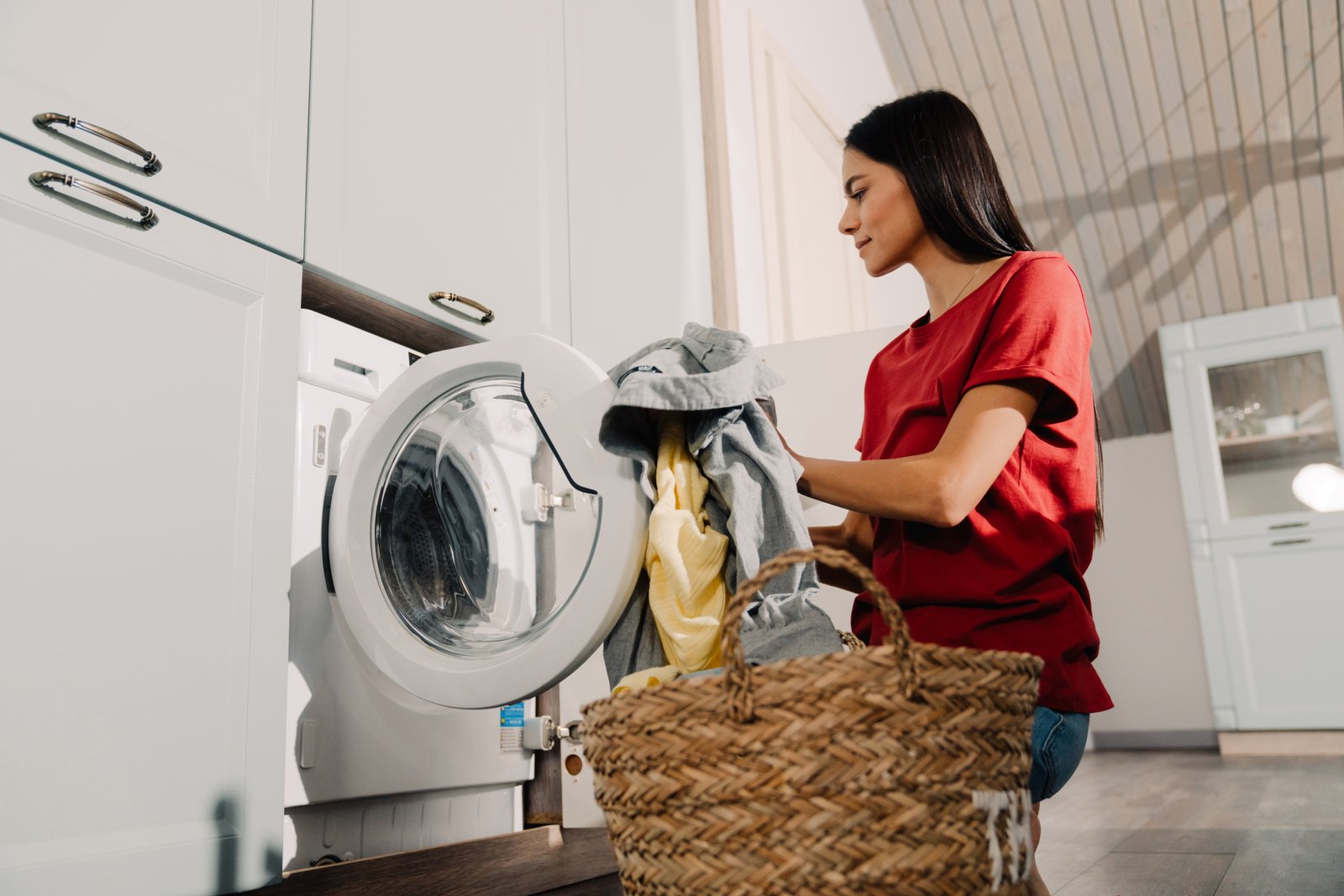 Young hispanic brunette woman putting clothes at washing machine while doing laundry at home